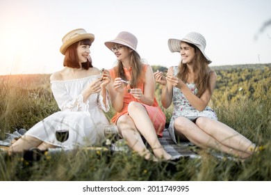 Female Friendship. Picnic Time. Three Attractive Girls Holding Escargot Snails In Hands And Eating, Against The Background Of Green Hills. Blanket With Food Spread On It.