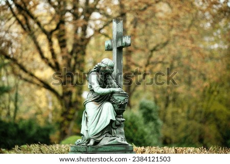 a female funerary figure sits mournfully at the foot of a cross with an urn against a blurred autumnal background