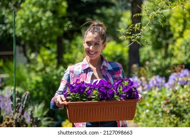 Female gardener holding rectangle planter looking to side smiling. Attractive girl holding potted violet petunias in garden. - Powered by Shutterstock