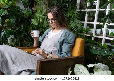 Female gardener wear linen dress, sitting on chair in green house, resting, using smartphone and drinking tea from a mug in harmony with the plants around. Home gardening, freelance. 