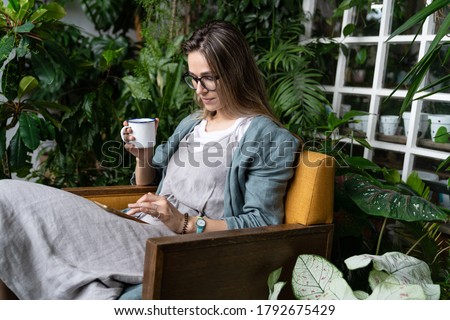 Female gardener wear linen dress, sitting on chair in green house, resting, using smartphone and drinking tea from a mug in harmony with the plants around. Home gardening, freelance. 