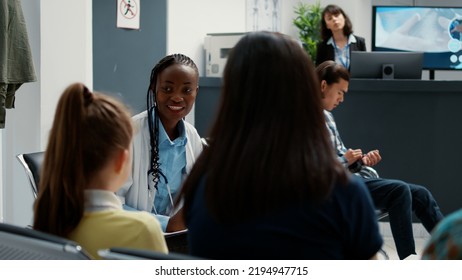 Female General Practitioner Talking To Mother With Little Girl For Consultation In Waiting Room Lobby. Medic Examining Woman And Child For Disease Treatment, Sitting In Hospital Reception.