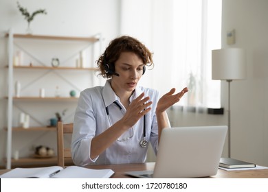 Female general practitioner wears white coat and headset speaking videoconferencing on laptop computer using online video call consultation app. Remote medical help for distance patient, telemedicine.