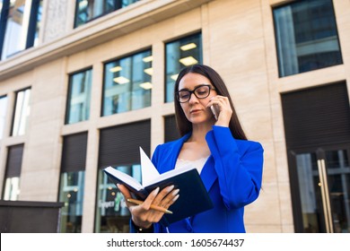 Female In Glasses Experienced Government Worker Reading Work Schedule From Diary And Talking Via Cell Telephone While Standing Outdoors Near Office Building 