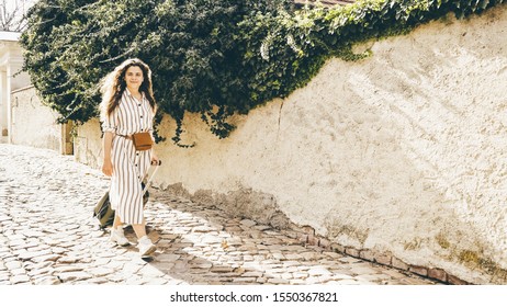 Female Going With Travel Luggage At The Narrow Street Between Residential Buildings In European Town. Young Girl Rolling Suitcase Down On The Cobblestone Pavement And Checking Route On The Paper Map