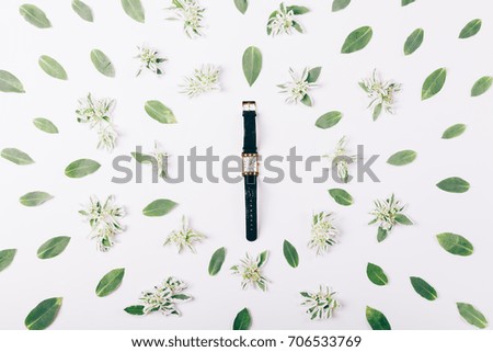 Female gold watch among green flowers and leaves on white table top view