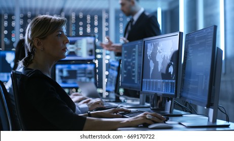 Female Government Employee Works In A Monitoring Room. In The Background Supervisor Holds Briefing. Possibly Government Agency Conducts Investigation. 