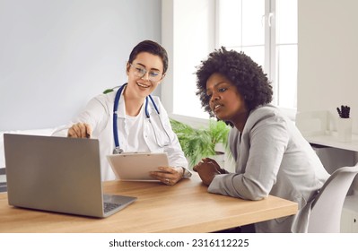 Female GP doctor showing test result to patient on laptop screen. Smiling female therapist giving advice to young African American woman. Patient visiting female doctor at clinic office - Powered by Shutterstock