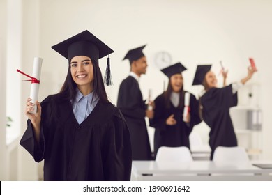 Female graduate dressed in a bachelor's gown shows the camera a diploma in her hands while standing in the classroom against the background of classmates who is taking a selfie. Education concept.