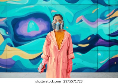 Female graffiti artist in respirator mask standing near the wall with her paintings looking to camera. Street art concept