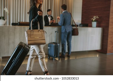 Female Guest Walks Inside A Hotel Lobby With People In The Background At Reception Counter. Woman Arriving At Hotel.