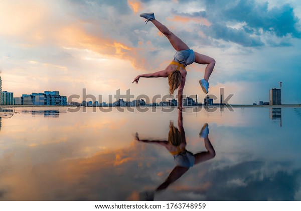Female gymnast\
standing on one hand and keeping balance during dramatic sunset\
with reflection in the water of amazing clouds. Concept of\
Calisthenic, contortion and handstand\
