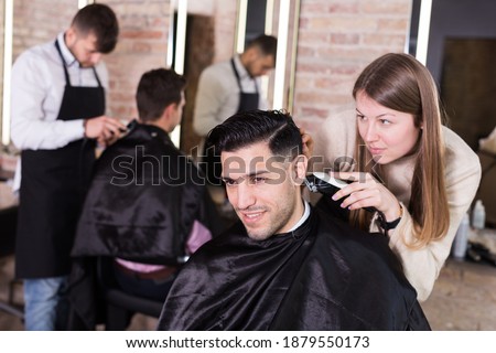 Female hairdresser cutting hair of male client with electric clipper in hair salon