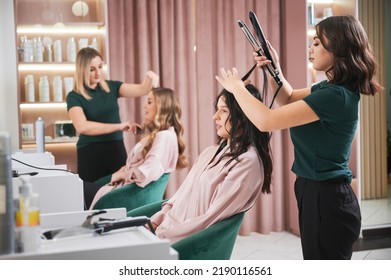 Female hairstylist curling client hair with curling iron device while colleague combing woman hair. Professional hairdresser doing hairstyle for woman in beauty salon. - Powered by Shutterstock