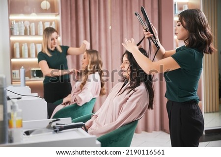 Female hairstylist curling client hair with curling iron device while colleague combing woman hair. Professional hairdresser doing hairstyle for woman in beauty salon.