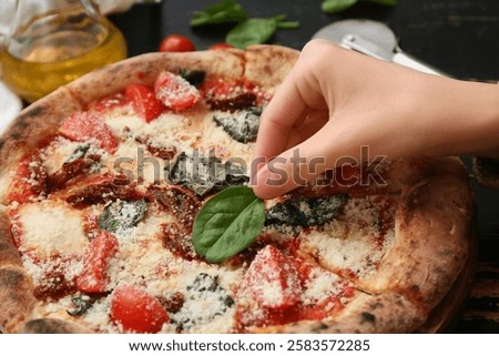 Female hand adding basil leaf to pizza Margarita on black wooden background. Closeup