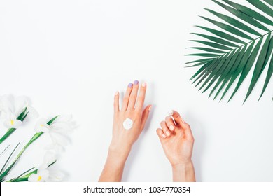 Female Hand Apply Cosmetic Cream On White Background Among Flowers, Top View