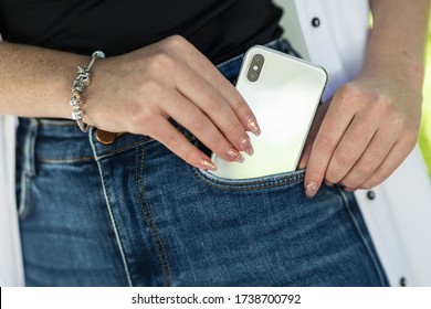 Female hand with beautiful bright design of manicure nails putting modern 5G smart phone in blue jeans pocket.