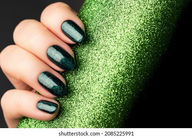 Female hand with beautiful holiday manicure - green glittered nails with glitter paper background with copy space. Selective focus. Nail care concept