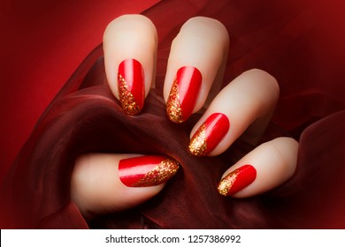 Female hand with beautiful red and golden glittered nails is holding a dark red textile on red background, manicure and nail care concept.