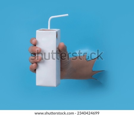 Female hand with blank packet carton juice  milk breaks through blue paper background.