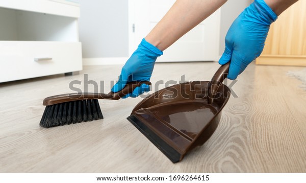 \
Female hand in a blue glove with\
a brush and dustpan on the floor background close-up. House\
cleaning concept. Cleaning microbes from bacteria and\
viruses.