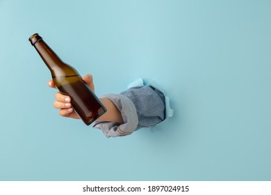 Female hand with bottle of beer breaks through blue paper background.