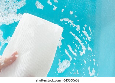 Female Hand Cleaning Window With Paper Towel And Detergent On Blue Background. Soap Stains On The Glass. Home Cleaning Concept.