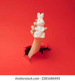 A female hand comes out from the red paper and holds white Easter bunny.