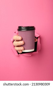 Female hand with cup of coffee punching through the pink paper. Top view