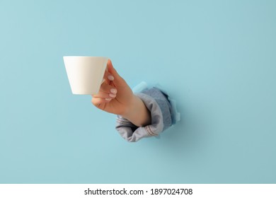 Female Hand With Cup Of Tea Or Coffee Breaks Through Blue Paper Background.