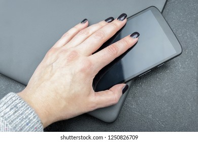 Female Hand With Dark Nails Holds A Mobile Phone, Which Lies On A Closed Gray Laptop