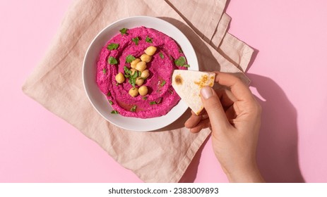 Female hand dipping pita bread triangles in bowl of beetroot hummus on pink background, top view, hard shadow