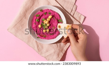 Female hand dipping pita bread triangles in bowl of beetroot hummus on pink background, top view, hard shadow
