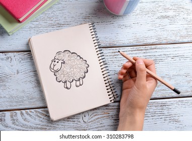 Female hand drawing sheep in notebook wooden table background