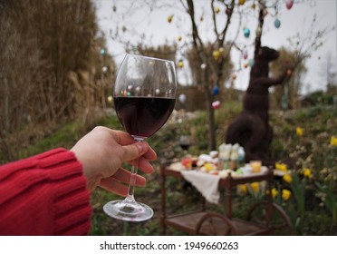 Female Hand With Glass Of Red Wine And Easter Tree With Decoration And Festive Breakfast Brunch On The Retro Mobile Table  In The Garden.