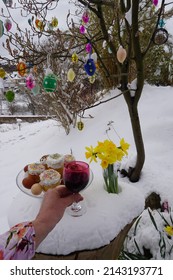 Female Hand With Glass Of Red Wine And Easter Tree With Decoration. Festive Breakfast Brunch On The Tree Trunk As Nature Table In The Snowy Garden.