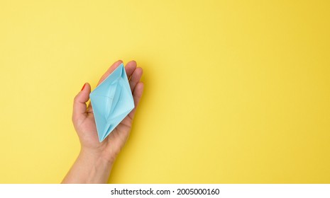 Female Hand Hold A Blue Paper Boat On A Yellow Background. Mentoring And Support Concept, Top View