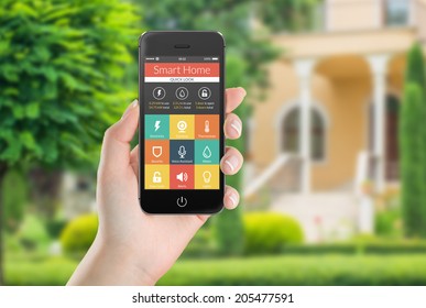 Female Hand Holding Black Mobile Smart Phone With Smart Home Application On The Screen. Blurred House On The Background. For Access To All Of The Controls Of Your House And Caring Of Home Security.