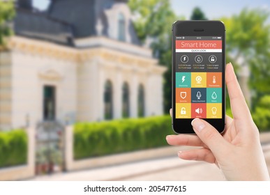 Female Hand Holding Black Mobile Smart Phone With Smart Home Application On The Screen. Blurred House On The Background. For Access To All Of The Controls Of Your House And Caring Of Home Security.