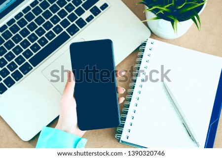 Female hand holding black mobile smart phone with blank screen on laptop computer and notepad background.