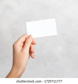 Female Hand Holding A Blank Business Card, Cutaway On Gray Concrete Background With Copy Space. Mockup