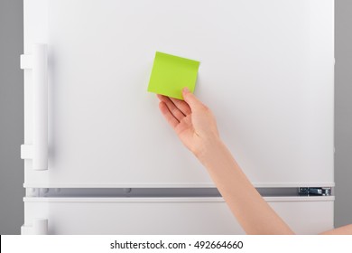 Female hand holding blank green sticky paper note on white refrigerator door