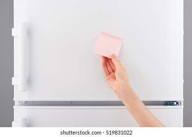 Female Hand Holding Blank Light Pink Sticky Paper Note On White Refrigerator Door