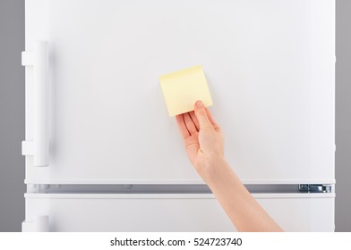 Female Hand Holding Blank Light Yellow Sticky Paper Note On White Refrigerator Door