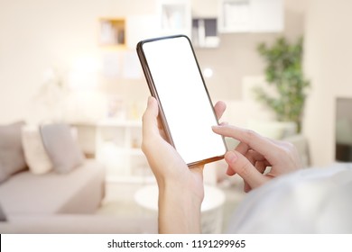 Female Hand Holding Blank Screen Mobile Smart Phone Over Living Room.