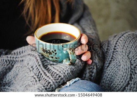 Female hand holding a blue cup of black coffee wrapped in warm knitted grey plaid. Autumn or Winter concept.