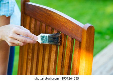 Female Hand Holding A Brush Applying Varnish Paint On A Wooden Garden Chair - Painting And Caring For Wood With Oil