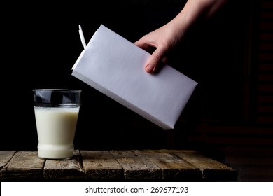 Female Hand Holding A Carton Of Milk And Pour Into A Glass On A Wooden Vintage Background