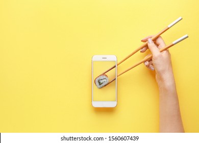 Female hand holding chopsticks and mobile phone with tasty sushi roll on screen against color background - Powered by Shutterstock